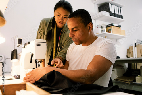 Female colleague assisting male tailor stitching clothes using sewing machine at workshop photo