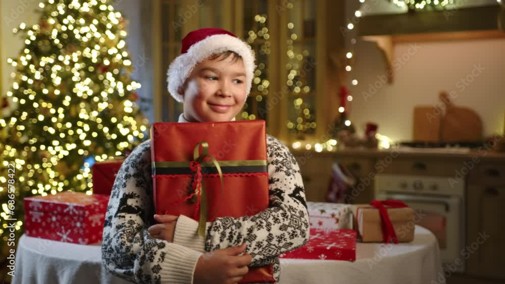 Dreamful 9-10 years old kid in warm ornamented sweater holding big box in red wrapping paper with ribbon. Beautiful Christmas tree with glowing garland. High quality 4k footage Dreamful 9-10 years old kid in warm ornamented sweater holding big box in red wrapping paper with ribbon. Beautiful Christmas tree with glowing garland. High quality 4k footage