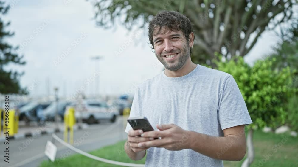 Happy young man, sporting a beard and blond hair, enjoying his time outdoor in the city, confidently smiling while typing a message on his smartphone screen Happy young man, sporting a beard and blond hair, enjoying his time outdoor in the city, confidently smiling while typing a message on his smartphone screen