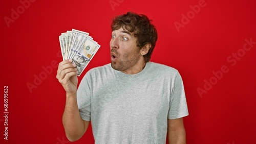 Shocked young man holding us banknotes, agape in amazement, fear, and disbelief against a stark red backdrop! wow, what a surprised face!
