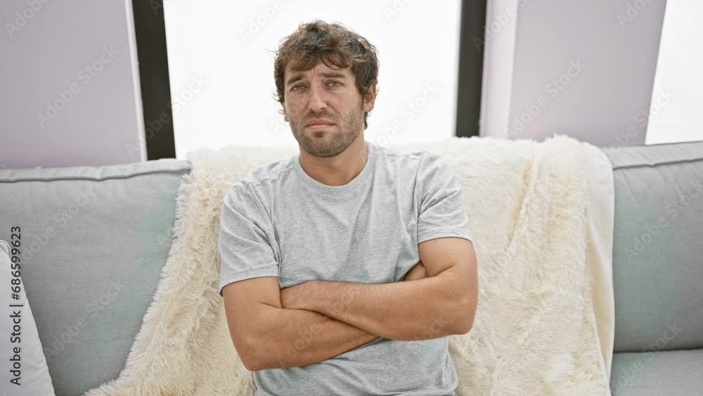 Skeptical young man with a disapproving expression, sitting on his cozy sofa, arms crossed, engulfed in negative emotions at home Skeptical young man with a disapproving expression, sitting on his cozy sofa, arms crossed, engulfed in negative emotions at home