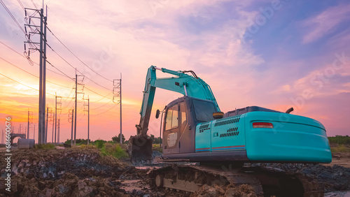 Excavator is leveling the ground in large yard for construction area of industrial building with row of electric poles against sunset sky in evening time