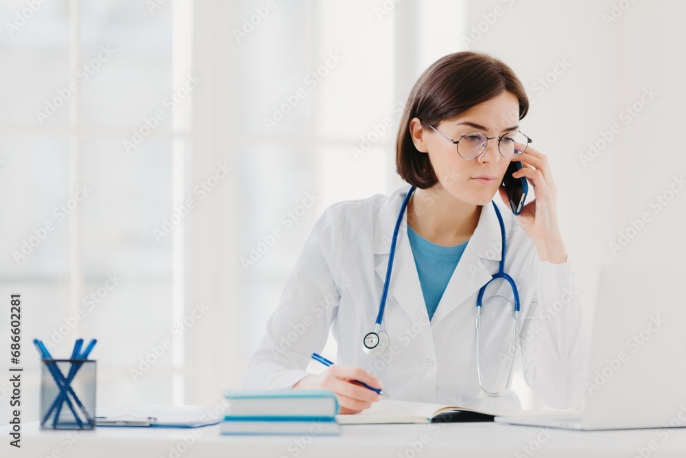 Focused female doctor on phone call with laptop and notes in office Focused female doctor on phone call with laptop and notes in office