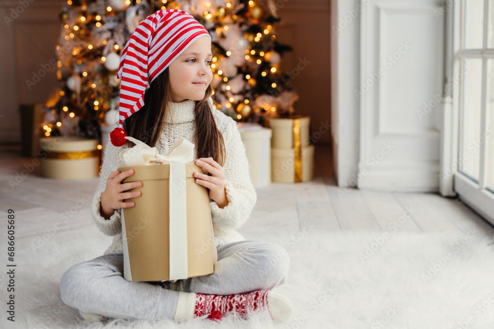 Fototapeta premium Girl in festive hat holding a Christmas gift, tree with golden lights behind