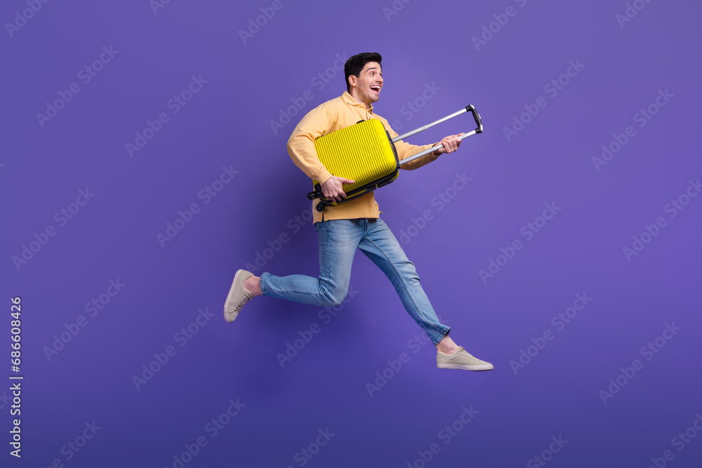 Full length photo of handsome young guy running hold baggage adventure wear trendy yellow garment isolated on violet color background Full length photo of handsome young guy running hold baggage adventure wear trendy yellow garment isolated on violet color background