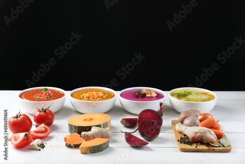 Tasty broth, different cream soups in bowls and ingredients on white wooden table against black background, space for text