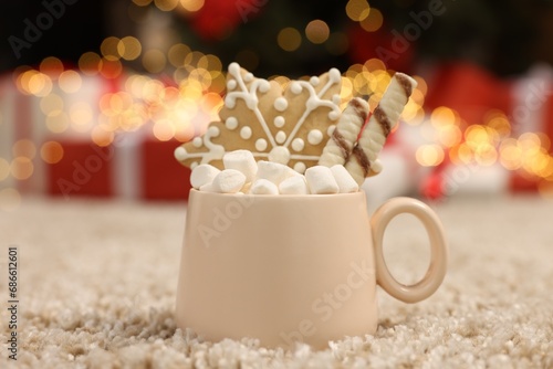 Delicious cocoa with marshmallows, wafer sticks and gingerbread cookie in cup on soft carpet against Christmas lights, closeup