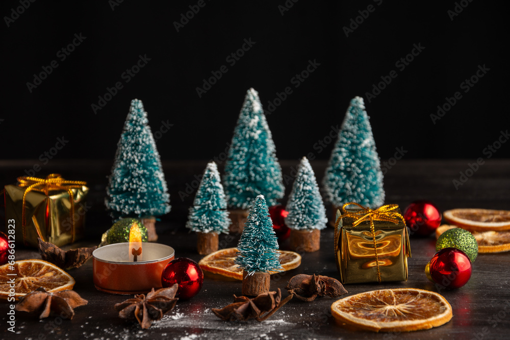 View of Christmas still life with snowy trees, balls, oranges, candle and gifts, black background, horizontal, with copy space