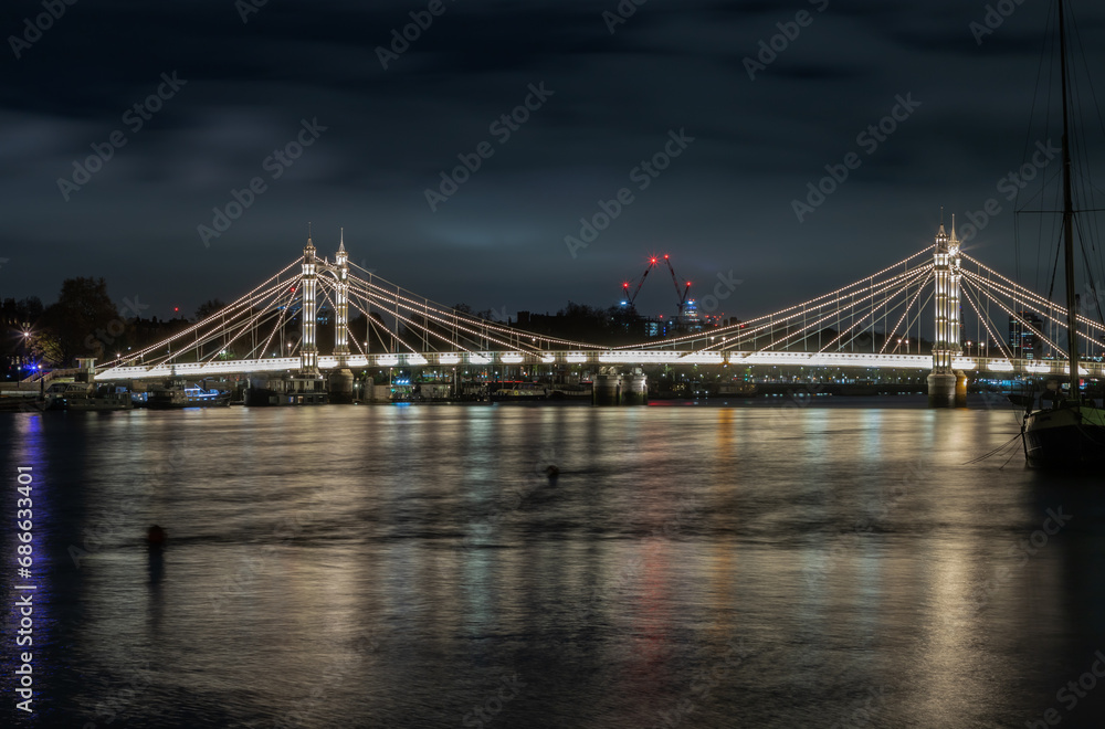 Fototapeta premium Nocturnal view of floodlit Albert Bridge from Chelsea Bridge with the reflection of the light in very calm water of River Thames. Lit up at Night Illuminated Albert bridge in west London at night. Spa