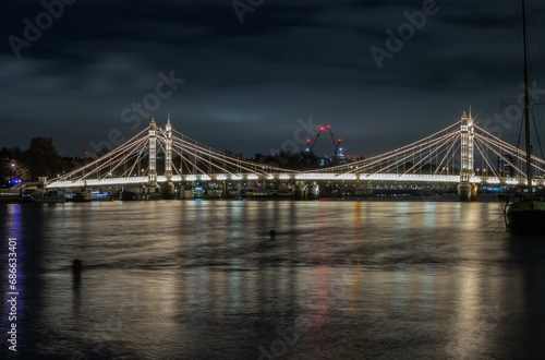 Nocturnal view of floodlit Albert Bridge from Chelsea Bridge with the reflection of the light in very calm water of River Thames. Lit up at Night Illuminated Albert bridge in west London at night. Spa