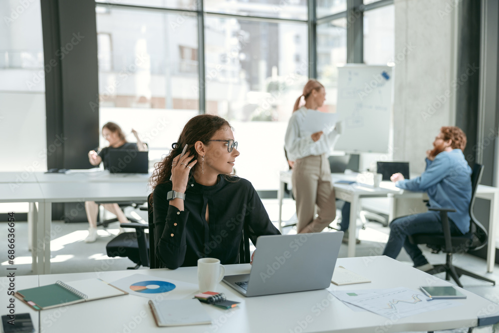 Smiling female entrepreneur working on laptop while sitting in modern coworking and looks away Smiling female entrepreneur working on laptop while sitting in modern coworking and looks away