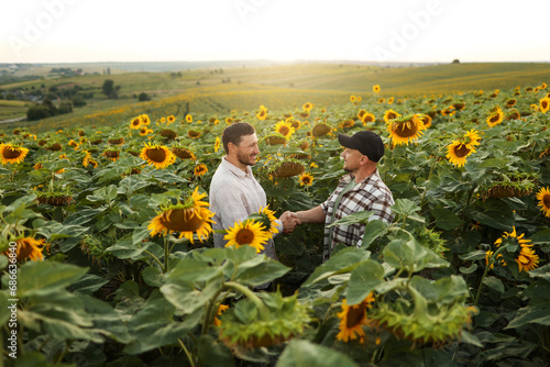 Agribusiness. Two smiling young farmers shaking hands and making advantageous business deal in blooming sunflowers field on summer. Teamwork of business partners. Concept of organic agriculture. photo