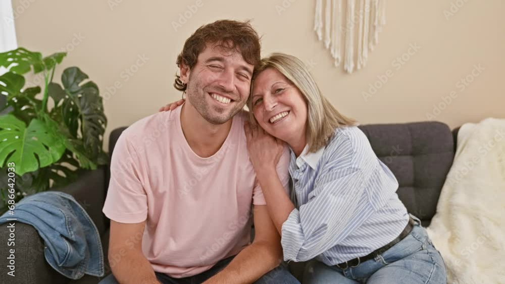 Heartwarming mother and son sitting together on the living room sofa, joyfully hugging and smiling in their cozy home, radiating happiness and confidence in their casual lifestyle. Heartwarming mother and son sitting together on the living room sofa, joyfully hugging and smiling in their cozy home, radiating happiness and confidence in their casual lifestyle.