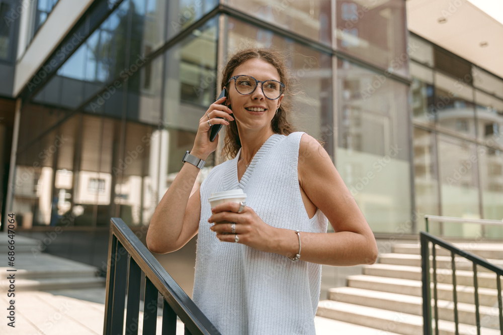 Pretty businesswoman with coffee cup standing on modern building background and talking phone