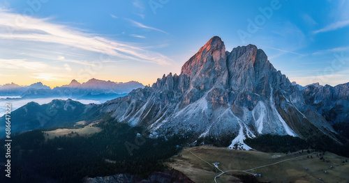 Sass de Puta mountain peak at Passo delle Erbe pass against the Dolomite peaks in the background, inverse cloud cover in the valley, blue sky, sunrise. Aerial drone view, large panorama. 