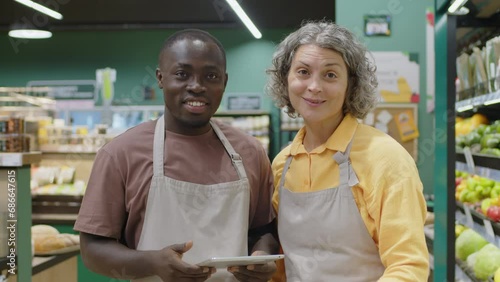 Medium portrait of two diverse male and female supermarket workers in aprons looking at digital tablet and having conversation then smiling at camera standing together in aisle with grocery photo