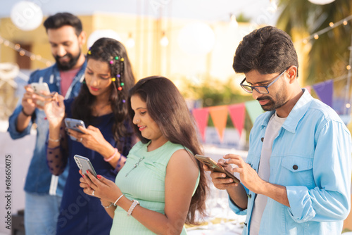 Group of young friends busy using mobile phones during birthday party or gathering on terrace - concept of millennial generation, social media sharing and cyberspace