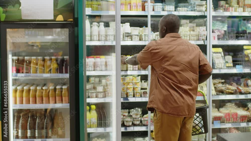 Medium shot of young African American man carrying shopping basket while standing by fridge with dairy products in supermarket aisle and choosing milk Medium shot of young African American man carrying shopping basket while standing by fridge with dairy products in supermarket aisle and choosing milk