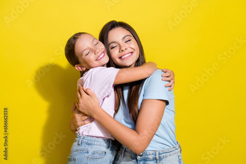 Photo of dreamy adorable little sisters wear t-shirts hugging closed eyes empty space isolated yellow color background