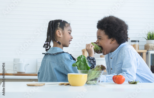 African little cute sister feeding smiling brother fresh green salad vegetable at home kitchen. Children having fun tasting vary vegetables together. Healthy food with kids