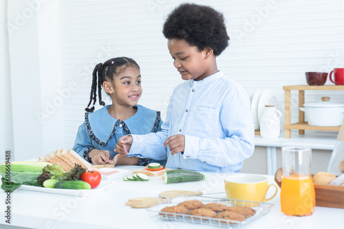 African children making sandwich, preparing food in kitchen together at home. Smiling brother and sister pick ham sliced, tomato, zucchini vegetables on top of bread. Orange juice and cookies on table