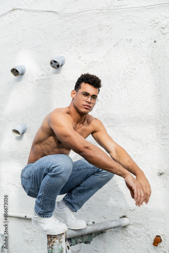 shirtless cuban man in stylish eyeglasses and blue jeans posing on urban street in Miami