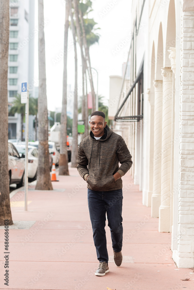 full length of cheerful african american man in hoodie and jeans walking with hands in pockets in Miami full length of cheerful african american man in hoodie and jeans walking with hands in pockets in Miami