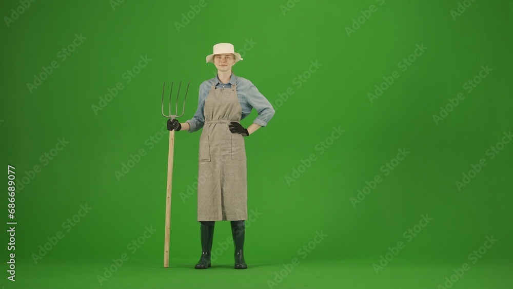 Portrait of farmer in working clothing on chroma key green screen. Gardener woman standing posing at camera with pitchfork, smiling face. Portrait of farmer in working clothing on chroma key green screen. Gardener woman standing posing at camera with pitchfork, smiling face.