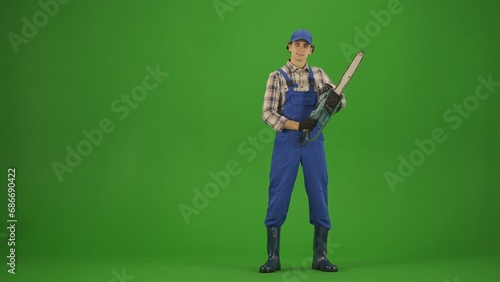 Portrait of farmer in working clothing on chroma key green screen. Gardener standing posing at the camera holding chainsaw.