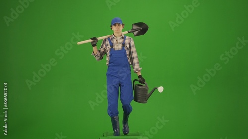 Portrait of farmer in working clothing on chroma key green screen. Gardener walking holding shovel over shoulder and watering can.