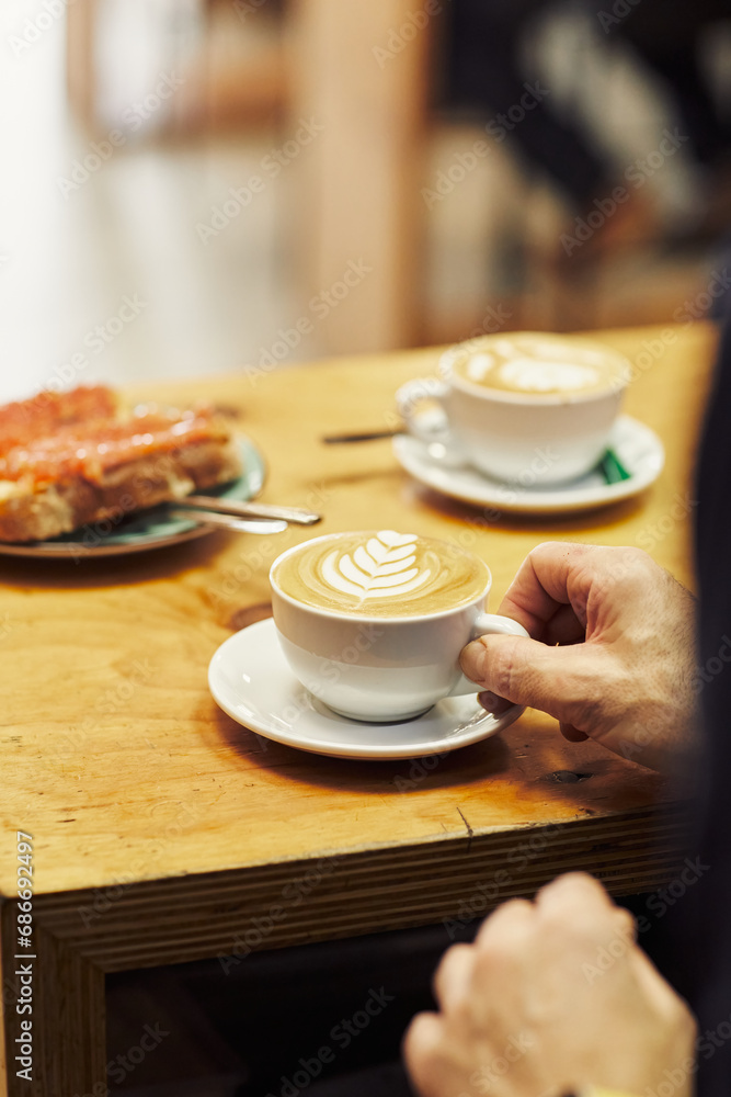 Hand reaching for a cappuccino with latte art on a wooden cafe table, another cup and plate in the background Hand reaching for a cappuccino with latte art on a wooden cafe table, another cup and plate in the background