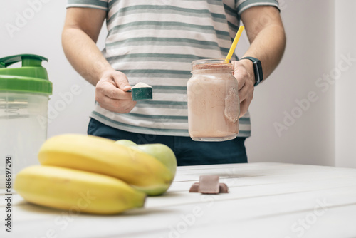 Man holding measuring spoon with protein powder, glass jar of protein drink cocktail, milkshake or smoothie above white wooden table with chocolate pieces, bananas, apples