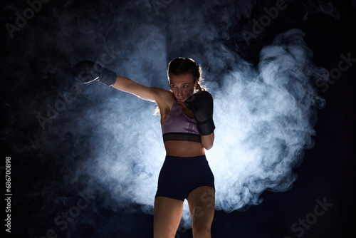 Dynamic portrait of sporty and powerful, young woman in boxing attire training in motion against black background in stage smoke. Concept of sport lifestyle, health.