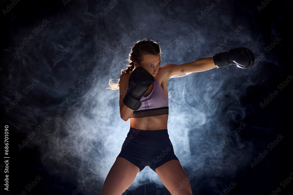 Dynamic and intense, female professional boxer showcases her athleticism against black studio background in stage smoke.