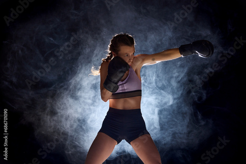 Dynamic and intense, female professional boxer showcases her athleticism against black studio background in stage smoke. © Lustre
