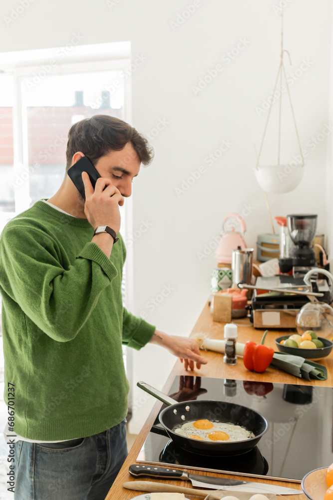 bearded man in jumper and jeans talking on smartphone while cooking breakfast in kitchen at home bearded man in jumper and jeans talking on smartphone while cooking breakfast in kitchen at home