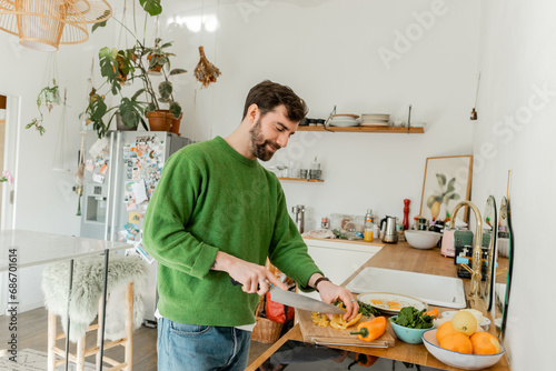 happy bearded man in jumper and jeans cutting fresh pepper while cooking breakfast in kitchen photo