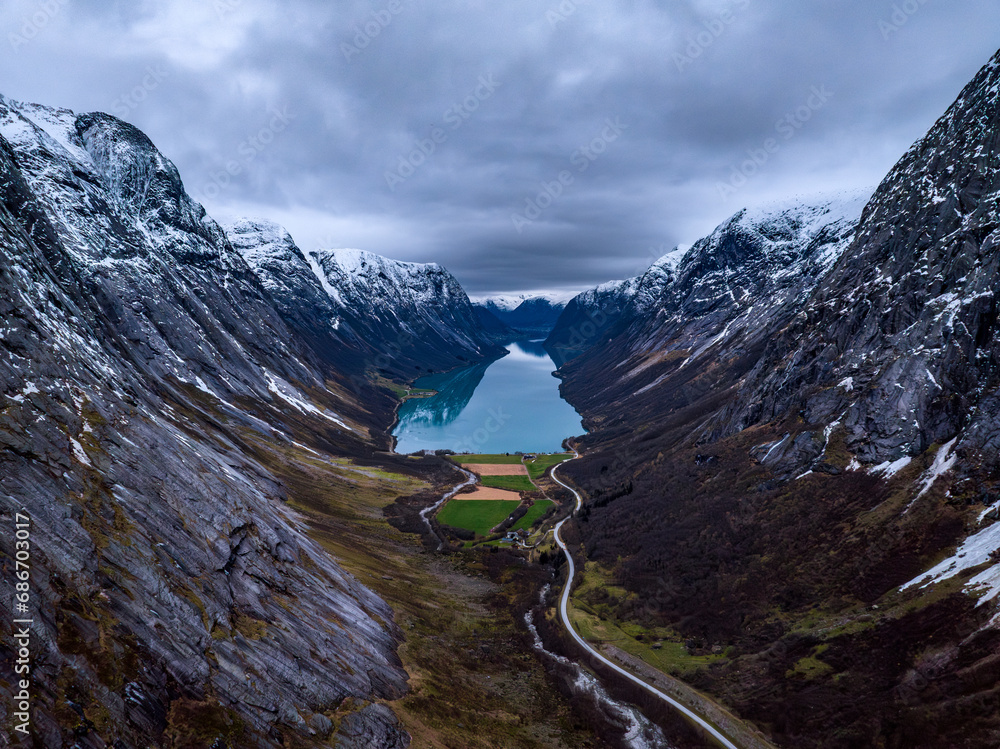 An aerial view of a Norwegian valley with symmetrically flanked mountains. where a farm and a mountain road are located, with a blue lake in the background.
