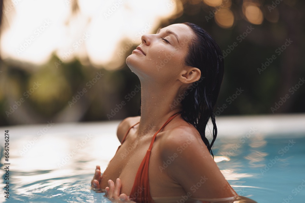 Tropical paradise, luxury relaxation of bikini brunette woman in sexy orange swimsuit in infinity pool at sunset covering her breast by hands. High quality portrait photo - obrazy, fototapety, plakaty 