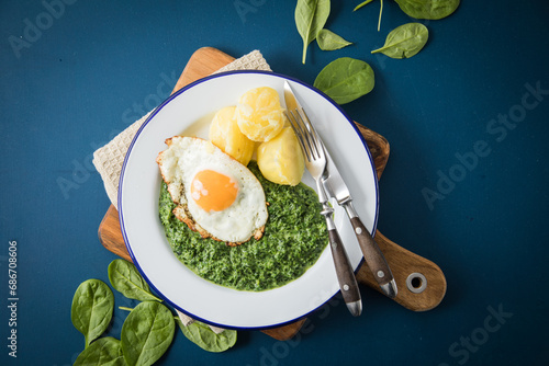 Rahmspinat mit Salzkartoffeln und Spiegelei auf Emaille Teller mit Besteck, Holzbrett, Serviette, Spinat Blätter roh und dunkel blau Hintergrund