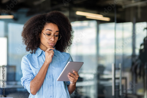 Serious thinking woman standing near window inside office, businesswoman using tablet computer, reading data with focus and concentration, female programmer in glasses and shirt testing programs