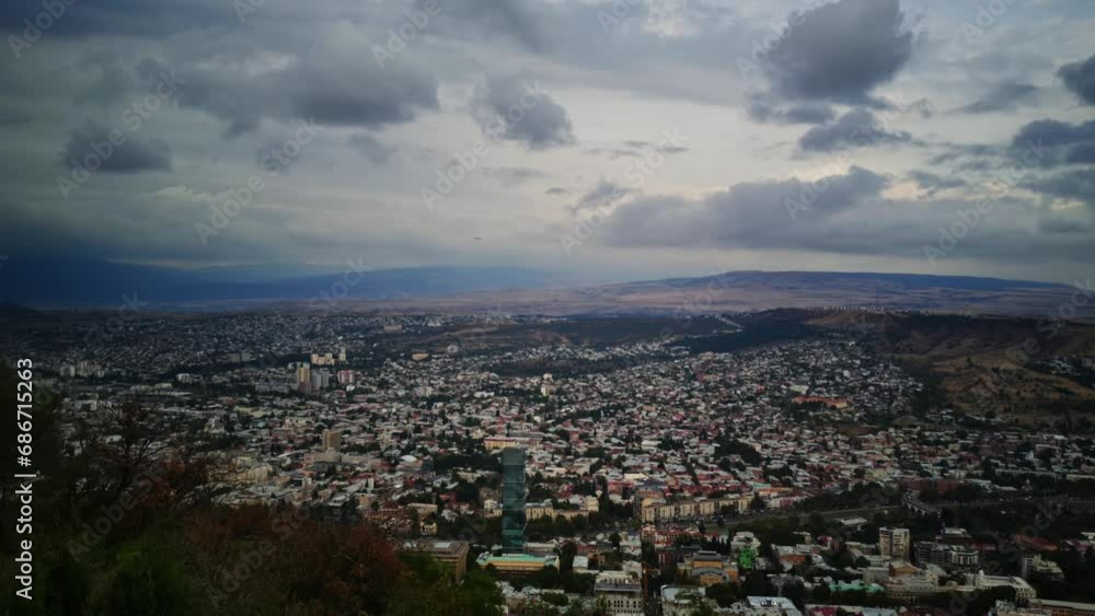 Tbilisi city filmed from high mountain with rocks range on the background. Scenery of Georgian town with streets full of houses and buildings under cloudy sky. Architecture and nature of Caucasus Tbilisi city filmed from high mountain with rocks range on the background. Scenery of Georgian town with streets full of houses and buildings under cloudy sky. Architecture and nature of Caucasus
