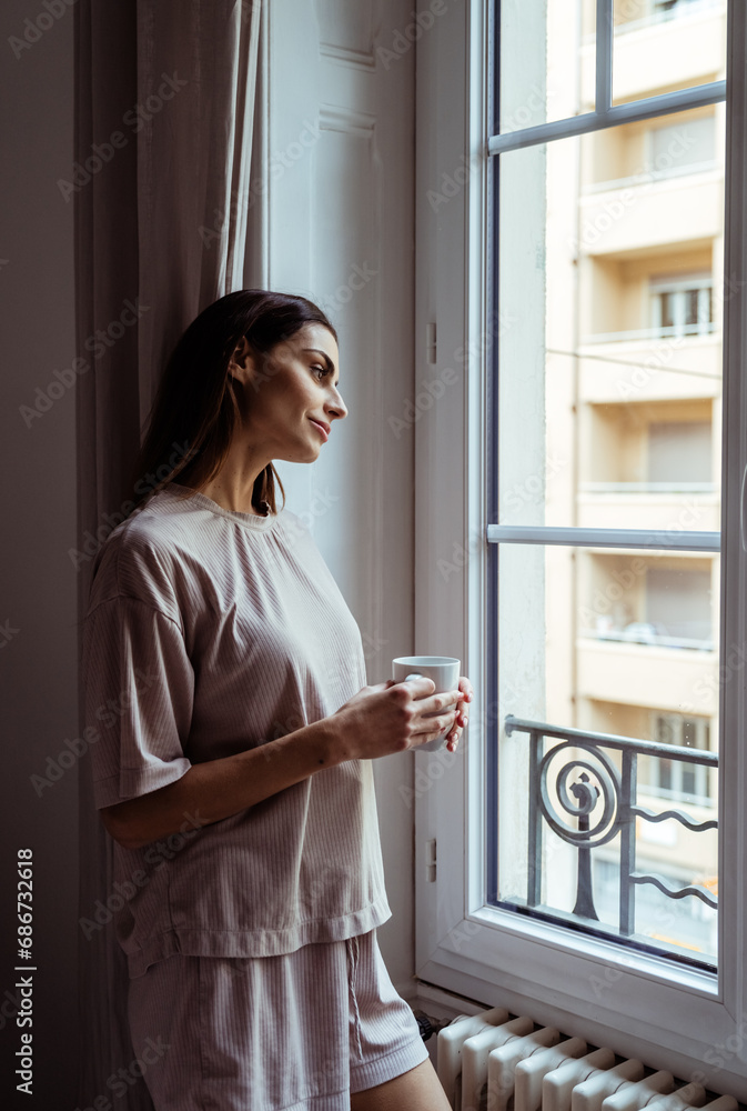 Beautiful thoughtful girl with long hair standing in bedroom looking at window drinking cofffee. Young woman feeling lonely looking at neighbor windows