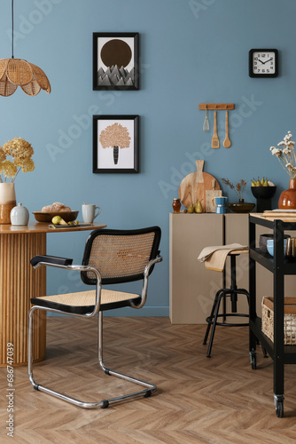 Warm and cozy dining room and kitchen interior with mock up poster frame, round table, rattan chair, beige commode, blue wall, vase with flowers and kitchen accessories. Home decor. Template.