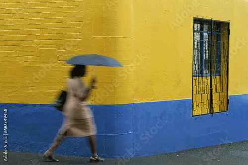 Woman runs past a yellow and blue wall holding an umbrella on a rainy day; Grenada, West Indies photo