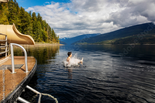 Boy splashing into the water after sliding out of the end of a houseboat waterslide on a fall day on Shuswap Lake; British Columbia, Canada photo