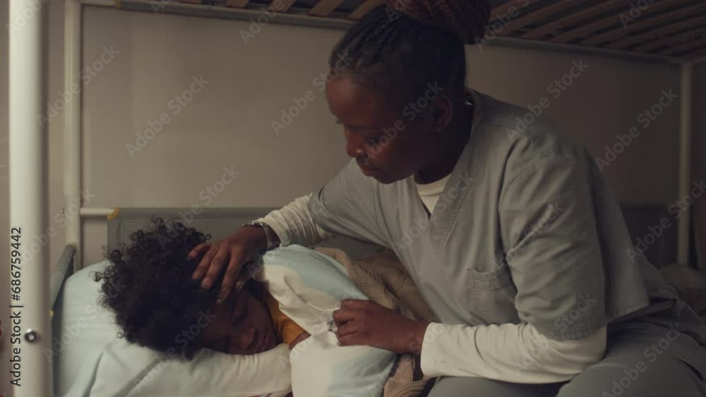 Medium shot of preteen African American boy sleeping in bedroom, mother in hospital gown walking in, tucking duvet, stroking his hair and kissing goodnight Medium shot of preteen African American boy sleeping in bedroom, mother in hospital gown walking in, tucking duvet, stroking his hair and kissing goodnight