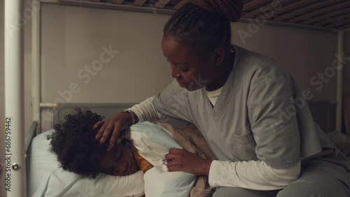 Medium shot of preteen African American boy sleeping in bedroom, mother in hospital gown walking in, tucking duvet, stroking his hair and kissing goodnight