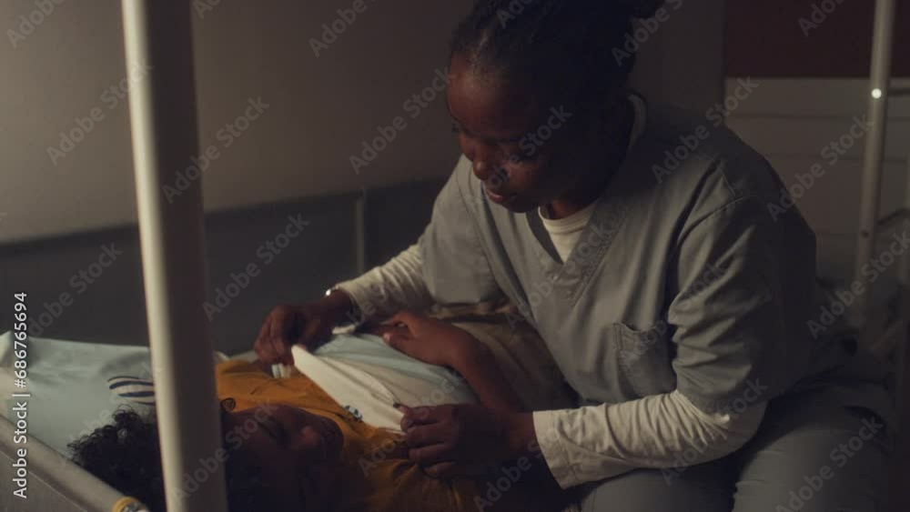 Medium pan shot of young black kid sleeping in bed at night, mother in health worker uniform coming in, tucking duvet, stroking hair, kissing him, then leaving and closing bedroom door Medium pan shot of young black kid sleeping in bed at night, mother in health worker uniform coming in, tucking duvet, stroking hair, kissing him, then leaving and closing bedroom door