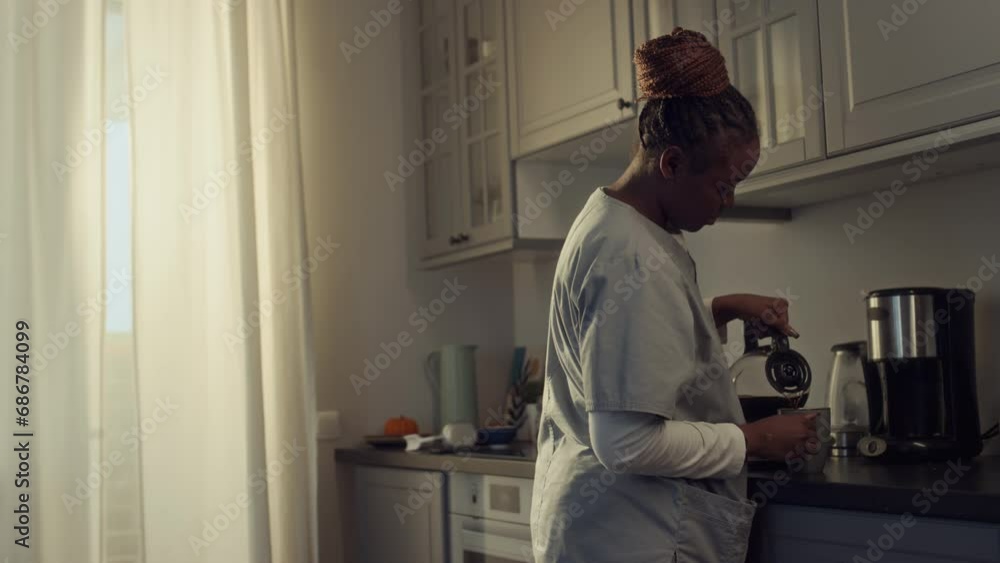 Medium shot of young African American woman in health worker uniform walking up to kitchen counter, pouring fresh coffee from coffee machine jug and drinking, while getting ready to go to work Medium shot of young African American woman in health worker uniform walking up to kitchen counter, pouring fresh coffee from coffee machine jug and drinking, while getting ready to go to work